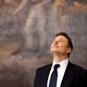 Elon Musk posa de terno e gravata olhando para cima durante evento, em imagem associada à sua fortuna recorde.