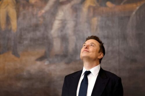 Elon Musk posa de terno e gravata olhando para cima durante evento, em imagem associada à sua fortuna recorde.