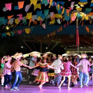 Quadrilha dança em festa junina com bandeirinhas coloridas e público acompanhando a celebração noturna.