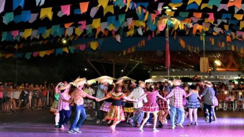 Quadrilha dança em festa junina com bandeirinhas coloridas e público acompanhando a celebração noturna.