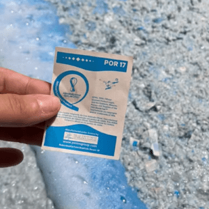 Uma pessoa segura com a mão esquerda o verso de uma figurinha oficial do álbum da Copa do Mundo do Catar de 2022, da Panini. A figurinha é retangular, de fundo branco com detalhes em azul, e traz no topo a inscrição "POR 17". No centro, há o logotipo oficial do torneio e textos informativos sobre direitos autorais e licenciamento em português. Na parte inferior, destaca-se uma faixa azul com o logotipo da Panini e o endereço do site da empresa. Ao fundo, a imagem mostra uma superfície texturizada e granulada em tons de azul-claro e branco, que se assemelha a uma grande quantidade de papel picado ou triturado.
