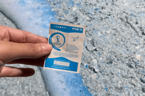 Uma pessoa segura com a mão esquerda o verso de uma figurinha oficial do álbum da Copa do Mundo do Catar de 2022, da Panini. A figurinha é retangular, de fundo branco com detalhes em azul, e traz no topo a inscrição "POR 17". No centro, há o logotipo oficial do torneio e textos informativos sobre direitos autorais e licenciamento em português. Na parte inferior, destaca-se uma faixa azul com o logotipo da Panini e o endereço do site da empresa. Ao fundo, a imagem mostra uma superfície texturizada e granulada em tons de azul-claro e branco, que se assemelha a uma grande quantidade de papel picado ou triturado.