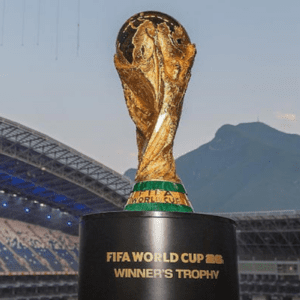 Uma imagem em primeiro plano do troféu da Copa do Mundo da FIFA, posicionado sobre uma base preta onde se lê "FIFA WORLD CUP 26 WINNER'S TROPHY". Ao fundo, vê-se a estrutura da arquibancada e da cobertura de um estádio de futebol, além de montanhas sob um céu claro de fim de tarde.