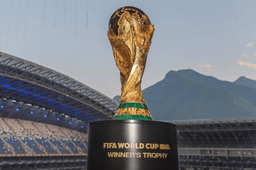 Uma imagem em primeiro plano do troféu da Copa do Mundo da FIFA, posicionado sobre uma base preta onde se lê "FIFA WORLD CUP 26 WINNER'S TROPHY". Ao fundo, vê-se a estrutura da arquibancada e da cobertura de um estádio de futebol, além de montanhas sob um céu claro de fim de tarde.