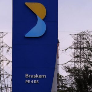 Totem da Braskem em frente a complexo petroquímico, com torres industriais e linhas de transmissão ao fundo.