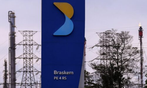 Totem da Braskem em frente a complexo petroquímico, com torres industriais e linhas de transmissão ao fundo.