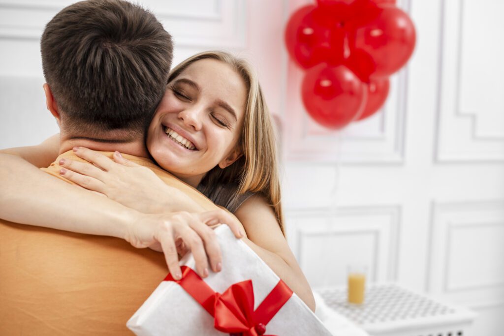 Uma mulher sorridente com os olhos fechados abraça um homem pelas costas. Ela segura um presente embrulhado em papel branco com um laço vermelho estruturado. O homem está de costas para a câmera, vestindo uma camiseta laranja. Ao fundo, o ambiente é claro e decorado com balões vermelhos e um copo de suco sobre uma mesa de cabeceira branca.