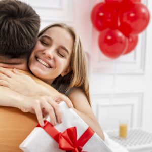 Uma mulher sorridente com os olhos fechados abraça um homem pelas costas. Ela segura um presente embrulhado em papel branco com um laço vermelho estruturado. O homem está de costas para a câmera, vestindo uma camiseta laranja. Ao fundo, o ambiente é claro e decorado com balões vermelhos e um copo de suco sobre uma mesa de cabeceira branca.