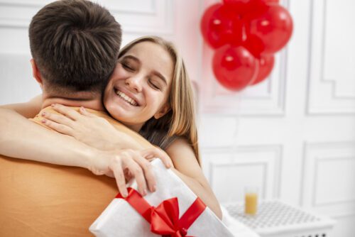 Uma mulher sorridente com os olhos fechados abraça um homem pelas costas. Ela segura um presente embrulhado em papel branco com um laço vermelho estruturado. O homem está de costas para a câmera, vestindo uma camiseta laranja. Ao fundo, o ambiente é claro e decorado com balões vermelhos e um copo de suco sobre uma mesa de cabeceira branca.