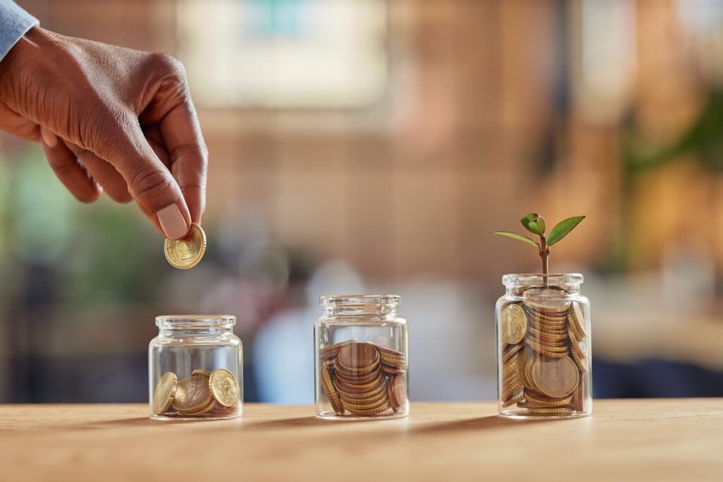 Mão colocando moeda em pote de vidro ao lado de outros com moedas e muda de planta, representando renda passiva.