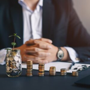 Pessoa ao fundo com pote de moedas e pilhas de moedas sobre a mesa, simbolizando investimentos e renda passiva.