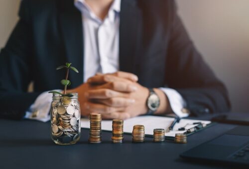 Pessoa ao fundo com pote de moedas e pilhas de moedas sobre a mesa, simbolizando investimentos e renda passiva.