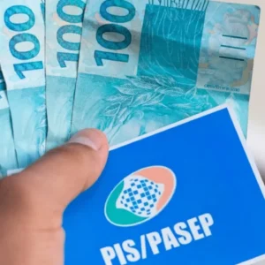 Mão segura cartão do PIS/Pasep e notas de R$ 100, representando pagamento do abono salarial a trabalhadores brasileiros.