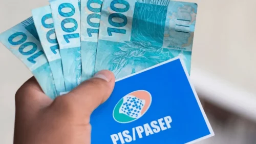Mão segura cartão do PIS/Pasep e notas de R$ 100, representando pagamento do abono salarial a trabalhadores brasileiros.