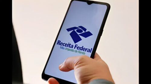 Pessoa segura celular com aplicativo Meu Imposto de Renda da Receita Federal aberto na tela.
