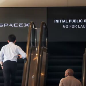Pessoas sobem escadas rolantes diante de painel da SpaceX anunciando a oferta pública inicial de ações da empresa.