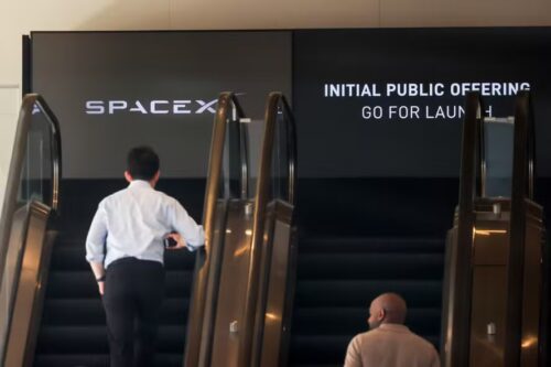 Pessoas sobem escadas rolantes diante de painel da SpaceX anunciando a oferta pública inicial de ações da empresa.