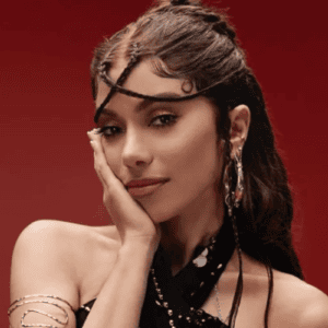 Retrato em plano médio da cantora Anitta contra um fundo vermelho sólido. Ela está levemente inclinada, apoiando o rosto delicadamente na mão direita. Seu cabelo castanho está estilizado de forma elaborada, com pequenas tranças finas que cruzam sua testa e o restante preso para trás. Ela usa uma maquiagem suave com sombra rosada brilhante, brincos prateados detalhados, um bracelete de metal espiralado no braço direito e uma blusa preta de frente única com detalhes em prata.