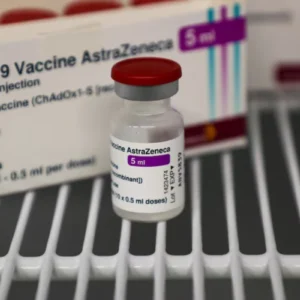 Fotografia focada em um frasco de vacina contra a COVID-19 da marca AstraZeneca, contendo tampa vermelha e rótulo com a inscrição '5 ml'. O frasco está sobre uma prateleira vazada de refrigerador e, ao fundo, aparece a caixa do medicamento.