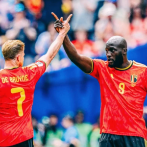 Fotografia em plano médio de dois jogadores de futebol da seleção da Bélgica comemorando em campo. À esquerda, de costas para a câmera, o jogador Kevin De Bruyne, vestindo a camisa vermelha número 7 com seu nome gravado em amarelo. À direita, de frente para a câmera, o jogador Romelu Lukaku, que é negro, tem a cabeça raspada, barba e veste a camisa vermelha número 9. Eles estão com os braços erguidos, tocando as mãos no alto em um gesto de celebração. O fundo mostra uma arquibancada lotada e desfocada em um estádio de futebol.