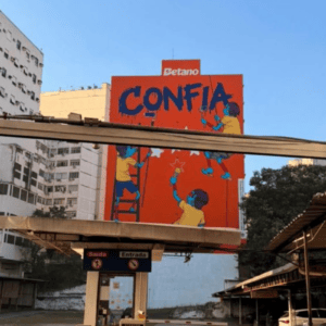 Uma fotografia em plano médio, tirada ao ar livre durante o dia, mostra um grande outdoor vertical de cor laranja vibrante no centro, posicionado atrás da entrada de um estacionamento. O outdoor exibe uma campanha da marca "Betano", com a palavra "CONFIA" pintada em grandes letras azuis de estilo grafite no topo. Abaixo da palavra, há uma ilustração estilizada de três figuras humanas de pele azul, vestindo camisas amarelas, que parecem estar pintando o outdoor: uma subindo uma escada à esquerda, outra suspensa por cordas à direita e uma terceira no centro, de costas, segurando um rolo de pintura. No topo do outdoor, acima da palavra "CONFIA", está o logotipo branco da "Betano". Em primeiro plano, vê-se a cobertura e as placas de "Saída" e "Entrada" do estacionamento. Ao fundo, à esquerda, há um grande edifício residencial ou comercial branco e antigo, e à direita, copas de árvores verdes e parte de um muro com pichações. O céu está limpo e azul claro.