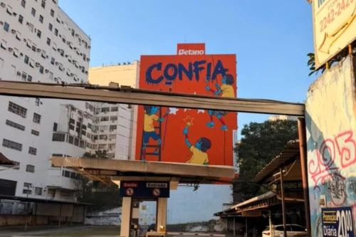 Uma fotografia em plano médio, tirada ao ar livre durante o dia, mostra um grande outdoor vertical de cor laranja vibrante no centro, posicionado atrás da entrada de um estacionamento. O outdoor exibe uma campanha da marca "Betano", com a palavra "CONFIA" pintada em grandes letras azuis de estilo grafite no topo. Abaixo da palavra, há uma ilustração estilizada de três figuras humanas de pele azul, vestindo camisas amarelas, que parecem estar pintando o outdoor: uma subindo uma escada à esquerda, outra suspensa por cordas à direita e uma terceira no centro, de costas, segurando um rolo de pintura. No topo do outdoor, acima da palavra "CONFIA", está o logotipo branco da "Betano". Em primeiro plano, vê-se a cobertura e as placas de "Saída" e "Entrada" do estacionamento. Ao fundo, à esquerda, há um grande edifício residencial ou comercial branco e antigo, e à direita, copas de árvores verdes e parte de um muro com pichações. O céu está limpo e azul claro.