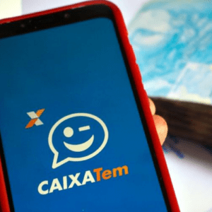 Uma pessoa segura um smartphone com uma capa vermelha, exibindo na tela azul o logotipo do aplicativo "CAIXA Tem", que traz um balão de fala com um rosto sorridente piscando e a logomarca da Caixa Econômica Federal. Ao fundo, fora de foco, há um maço de cédulas de 100 reais (reais brasileiros) sobre uma superfície branca.