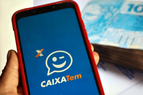 Uma pessoa segura um smartphone com uma capa vermelha, exibindo na tela azul o logotipo do aplicativo "CAIXA Tem", que traz um balão de fala com um rosto sorridente piscando e a logomarca da Caixa Econômica Federal. Ao fundo, fora de foco, há um maço de cédulas de 100 reais (reais brasileiros) sobre uma superfície branca.