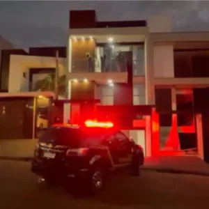 Fotografia noturna mostrando a fachada de uma casa moderna de alto padrão com vários andares e iluminação embutida. Em frente à residência, está estacionada uma viatura policial com os giroflex vermelhos acesos, iluminando a rua e a fachada do imóvel.