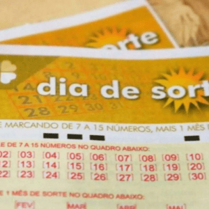 Foto em plano fechado e focada na parte superior de um volante de apostas da loteria brasileira "Dia de Sorte". O bilhete é predominantemente amarelo e branco. No topo, lê-se o nome do jogo "dia de sorte" em letras brancas sobre um fundo amarelo, acompanhado pelo desenho de um sol à direita e um trevo de quatro folhas à esquerda. Logo abaixo, há instruções em letras vermelhas e um quadro numérico que vai do 01 ao 31, organizado em linhas. Ao fundo, de forma desfocada, é possível ver outro volante igual.