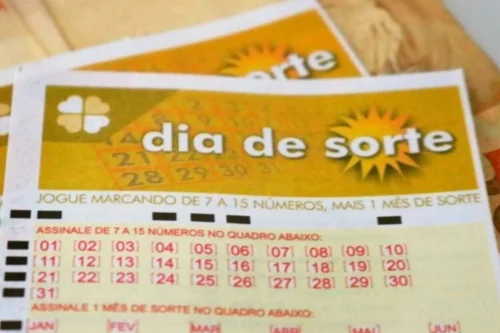 Foto em plano fechado e focada na parte superior de um volante de apostas da loteria brasileira "Dia de Sorte". O bilhete é predominantemente amarelo e branco. No topo, lê-se o nome do jogo "dia de sorte" em letras brancas sobre um fundo amarelo, acompanhado pelo desenho de um sol à direita e um trevo de quatro folhas à esquerda. Logo abaixo, há instruções em letras vermelhas e um quadro numérico que vai do 01 ao 31, organizado em linhas. Ao fundo, de forma desfocada, é possível ver outro volante igual.