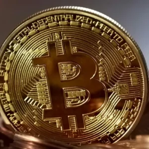 Uma moeda física dourada e brilhante com o símbolo estilizado do Bitcoin em destaque no centro. A moeda está em pé, apoiada sobre uma pilha de outras moedas de tom cobre, com um fundo levemente desfocado em tons cinza e marrom.