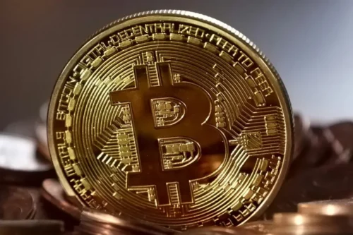 Uma moeda física dourada e brilhante com o símbolo estilizado do Bitcoin em destaque no centro. A moeda está em pé, apoiada sobre uma pilha de outras moedas de tom cobre, com um fundo levemente desfocado em tons cinza e marrom.