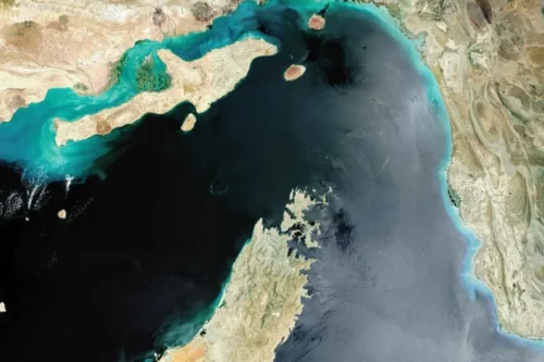 Imagem de satélite do Estreito de Ormuz, mostrando a Península de Musandam ao sul e a costa do Irã ao norte, com águas em tons de azul-escuro e turquesa que conectam o Golfo Pérsico ao Golfo de Omã.
