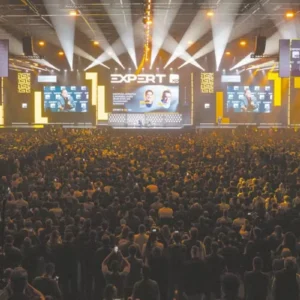 Vista traseira de uma grande multidão lotando um auditório iluminado durante o evento "Expert XP". No palco ao fundo, há painéis eletrônicos gigantes exibindo a transmissão ao vivo do palestrante e gráficos do evento, acompanhados por holofotes e iluminação amarela e preta.