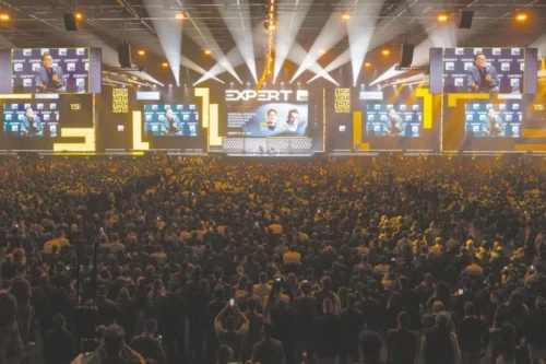 Vista traseira de uma grande multidão lotando um auditório iluminado durante o evento "Expert XP". No palco ao fundo, há painéis eletrônicos gigantes exibindo a transmissão ao vivo do palestrante e gráficos do evento, acompanhados por holofotes e iluminação amarela e preta.