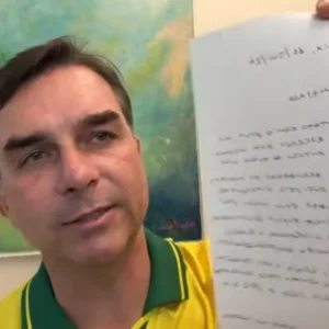 Close do senador Flávio Bolsonaro, vestindo uma camisa amarela e verde, enquanto segura uma folha de papel com um texto manuscrito em frente à câmera. O texto no papel está invertido (efeito espelho). Ao fundo, há uma pintura abstrata colorida.