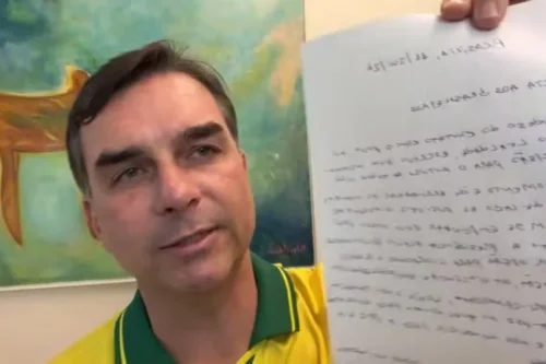 Close do senador Flávio Bolsonaro, vestindo uma camisa amarela e verde, enquanto segura uma folha de papel com um texto manuscrito em frente à câmera. O texto no papel está invertido (efeito espelho). Ao fundo, há uma pintura abstrata colorida.