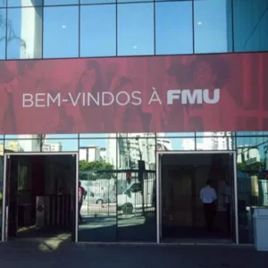 Fotografia da entrada de um campus universitário da FMU. A fachada possui painéis de vidro espelhado que refletem a rua e prédios ao redor. Logo acima das portas de entrada, há uma grande faixa vermelha com a frase "BEM-VINDOS À FMU" escrita em letras brancas sobrepostas a uma imagem suave de estudantes sorrindo. Nas laterais, veem-se lixeiras cinzas e detalhes do saguão de entrada através dos vidros.