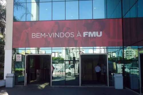 Fotografia da entrada de um campus universitário da FMU. A fachada possui painéis de vidro espelhado que refletem a rua e prédios ao redor. Logo acima das portas de entrada, há uma grande faixa vermelha com a frase "BEM-VINDOS À FMU" escrita em letras brancas sobrepostas a uma imagem suave de estudantes sorrindo. Nas laterais, veem-se lixeiras cinzas e detalhes do saguão de entrada através dos vidros.