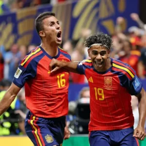 Dois jogadores da seleção espanhola de futebol celebram um gol em campo durante uma partida. À esquerda, o jogador de número 16 (Rodri) grita com entusiasmo, com o punho cerrado e os braços abertos. À direita, o jogador de número 19 (Lamine Yamal), sorrindo amplamente e usando uma faixa branca na testa, abraça o companheiro pelo peito. Ambos vestem o uniforme vermelho e azul da Espanha. Ao fundo, as arquibancadas do estádio aparecem desfocadas com torcedores comemorando.