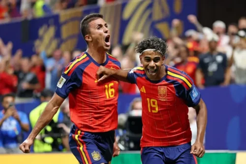 Dois jogadores da seleção espanhola de futebol celebram um gol em campo durante uma partida. À esquerda, o jogador de número 16 (Rodri) grita com entusiasmo, com o punho cerrado e os braços abertos. À direita, o jogador de número 19 (Lamine Yamal), sorrindo amplamente e usando uma faixa branca na testa, abraça o companheiro pelo peito. Ambos vestem o uniforme vermelho e azul da Espanha. Ao fundo, as arquibancadas do estádio aparecem desfocadas com torcedores comemorando.