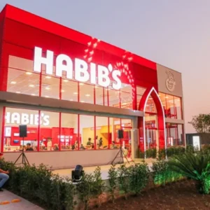 Fotografia da fachada de uma filial do Habib's ao entardecer. O prédio possui grandes janelas de vidro, estrutura vermelha e o letreiro luminoso da marca em branco no topo. É possível ver pessoas no interior do restaurante e vegetação paisagística na frente.