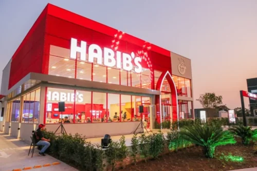 Fotografia da fachada de uma filial do Habib's ao entardecer. O prédio possui grandes janelas de vidro, estrutura vermelha e o letreiro luminoso da marca em branco no topo. É possível ver pessoas no interior do restaurante e vegetação paisagística na frente.