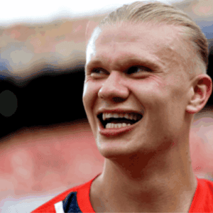 Retrato em close-up do jogador de futebol Erling Haaland sorrindo de forma descontraída e olhando para o lado. Ele tem cabelos loiros bem claros penteados e presos para trás. Ele veste uma camisa esportiva vermelha com detalhes em azul e branco na gola. O fundo da imagem está desfocado, mostrando a estrutura e as arquibancadas coloridas de um estádio de futebol sob a luz do dia.