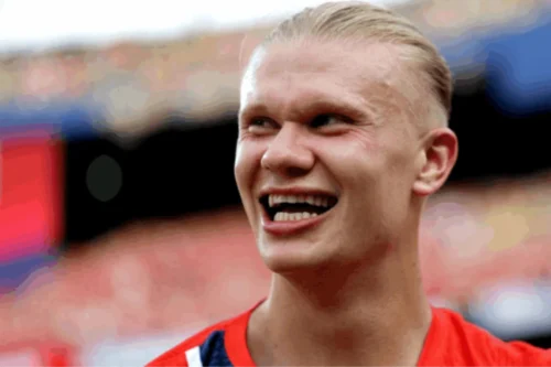 Retrato em close-up do jogador de futebol Erling Haaland sorrindo de forma descontraída e olhando para o lado. Ele tem cabelos loiros bem claros penteados e presos para trás. Ele veste uma camisa esportiva vermelha com detalhes em azul e branco na gola. O fundo da imagem está desfocado, mostrando a estrutura e as arquibancadas coloridas de um estádio de futebol sob a luz do dia.
