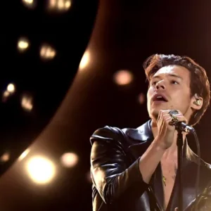 Fotografia de meio-corpo do cantor Harry Styles no palco durante uma apresentação. Ele está de perfil, olhando para cima e para a esquerda com a boca levemente aberta, segurando um microfone com a mão direita cheia de anéis chamativos. Ele veste uma jaqueta de couro preta brilhante. O fundo está escuro, iluminado por luzes de palco desfocadas em tons quocientes e dourados.