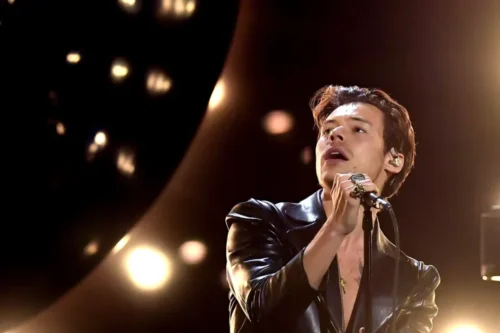 Fotografia de meio-corpo do cantor Harry Styles no palco durante uma apresentação. Ele está de perfil, olhando para cima e para a esquerda com a boca levemente aberta, segurando um microfone com a mão direita cheia de anéis chamativos. Ele veste uma jaqueta de couro preta brilhante. O fundo está escuro, iluminado por luzes de palco desfocadas em tons quocientes e dourados.