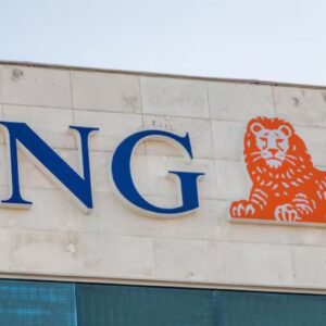 Foto em plano fechado da fachada de um edifício exibindo o logotipo da instituição financeira ING. À esquerda, destacam-se as letras "ING" em caixa alta, com uma fonte serifada na cor azul-escura e bordas brancas em relevo. À direita do texto, há o símbolo da marca: a ilustração estilizada de um leão deitado na cor laranja vibrante, também com contorno branco em relevo. O letreiro está fixado em uma parede de placas de concreto ou pedra clara, e logo abaixo é possível ver o topo de uma janela com vidros reflexivos azulados.