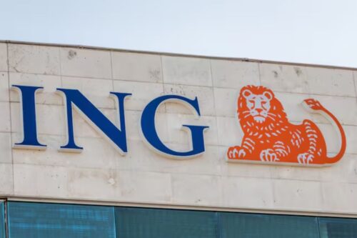 Foto em plano fechado da fachada de um edifício exibindo o logotipo da instituição financeira ING. À esquerda, destacam-se as letras "ING" em caixa alta, com uma fonte serifada na cor azul-escura e bordas brancas em relevo. À direita do texto, há o símbolo da marca: a ilustração estilizada de um leão deitado na cor laranja vibrante, também com contorno branco em relevo. O letreiro está fixado em uma parede de placas de concreto ou pedra clara, e logo abaixo é possível ver o topo de uma janela com vidros reflexivos azulados.